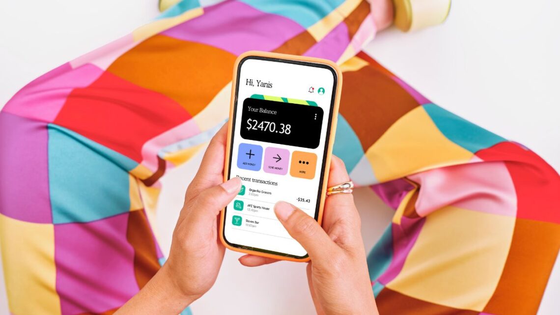 Osoba drží smartphone s otvorenou aplikáciou na správu financií, pričom na displeji je zobrazený zostatok $2470.38, obklopená farebným oblečením s geometrickým vzorom.