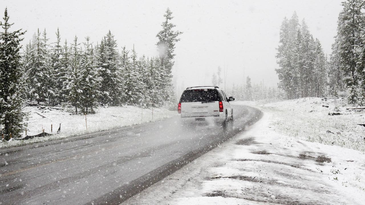 Auto jazdí po zasneženej ceste obklopenej stromami počas sneženia, ktoré pokrýva krajinu prvým snehom.