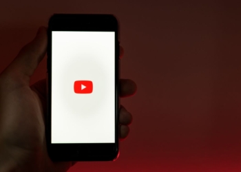 Ruka držiaca mobilný telefón s logom YouTube na obrazovke, s tmavým pozadím.