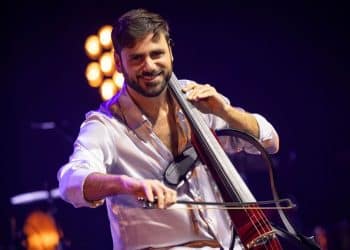 toznámy violončelista HAUSER, člen dua 2Cellos, hrá na elektrickom violončele počas koncertu, oblečený v bielej košeli, s úsmevom na tvári, v osvetlenej koncertnej sále.